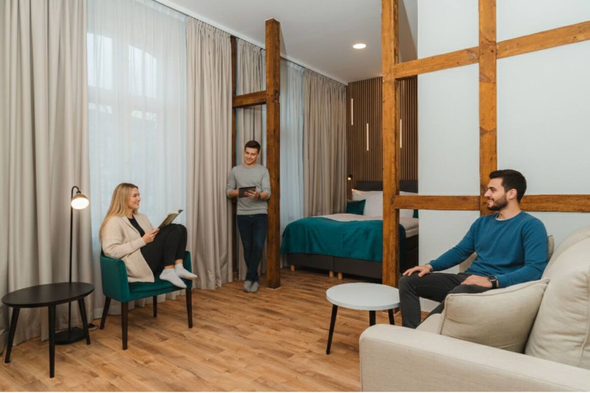 Ein modernes Hotelzimmer mit Holzboden, einem Bett mit grüner Bettwäsche, einer Couch, einem kleinen Tisch und einer Stehlampe.