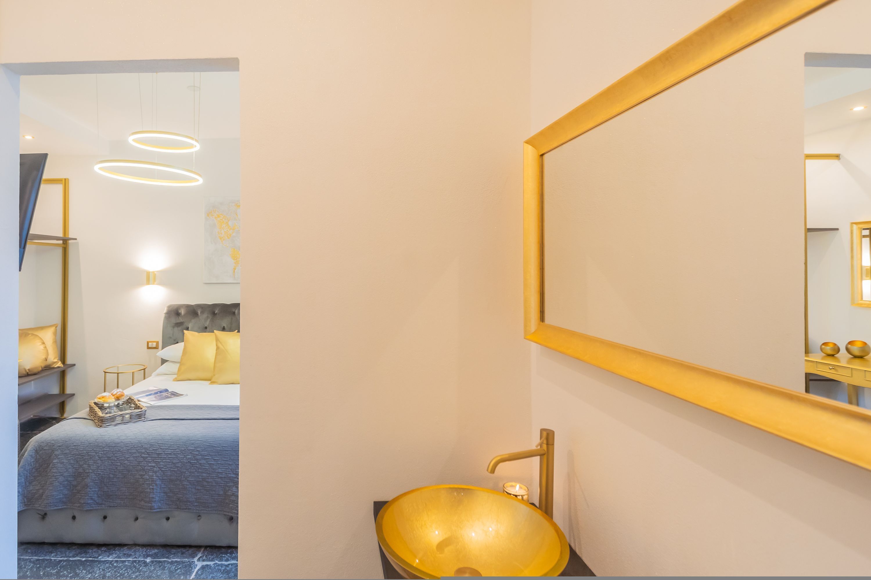 Un baño moderno con un espejo grande, un lavabo dorado y una cama con cabecera gris y almohadas amarillas.