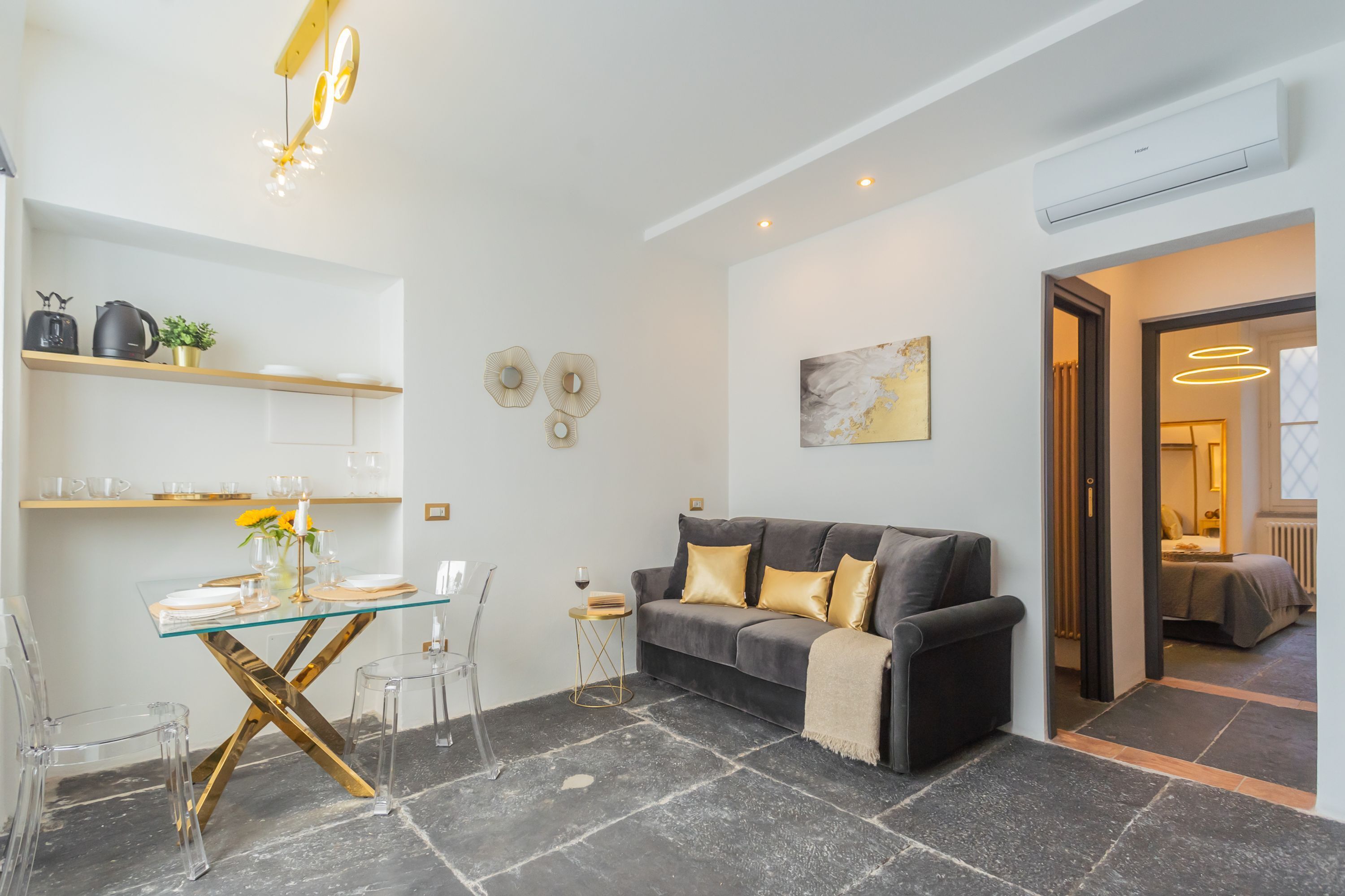 A modern living room with a glass dining table, a sofa with cushions, a coffee machine, and a small side table.