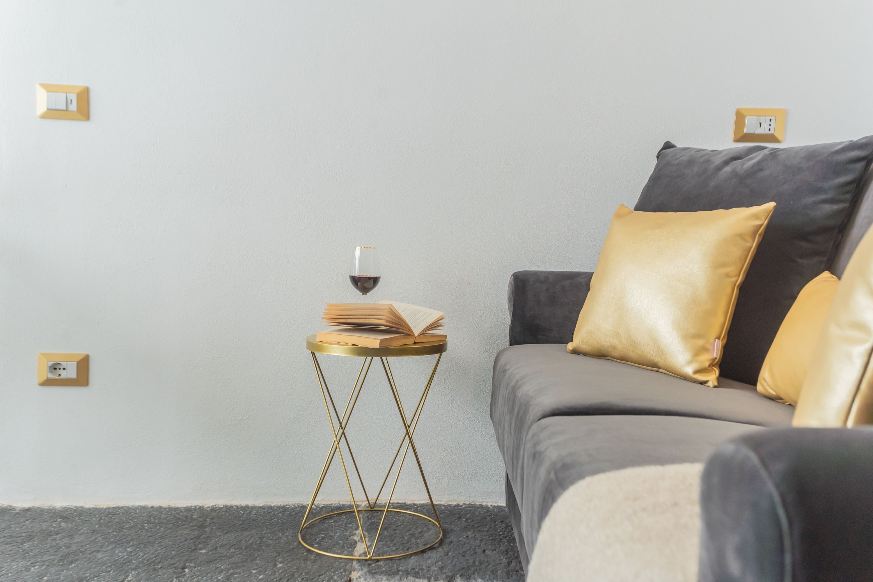 A modern living room with a grey sofa, gold cushions, and a small round side table with a book and a glass of wine.