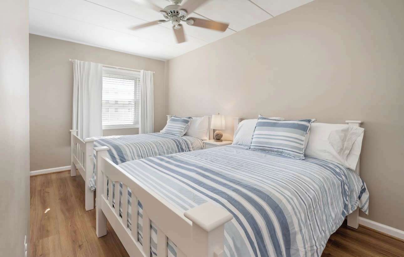 A bedroom with two beds, a ceiling fan, a window with curtains, and a bedside lamp.