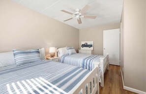 Un dormitorio con dos camas, un ventilador de techo, una mesita de noche con lámpara y una puerta que da a otra habitación.