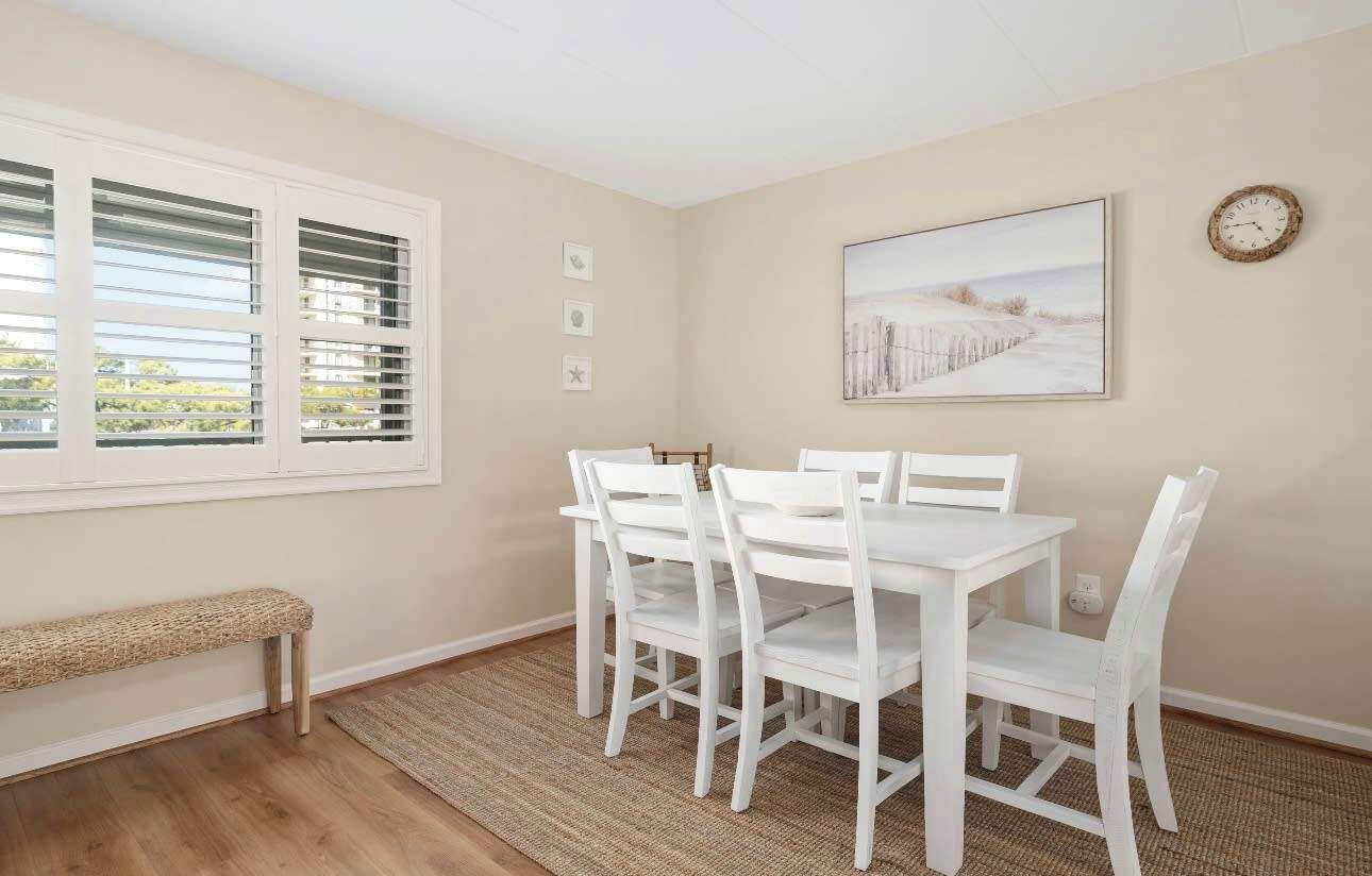 A dining area with a white table and chairs, a bench, and a window with blinds.
