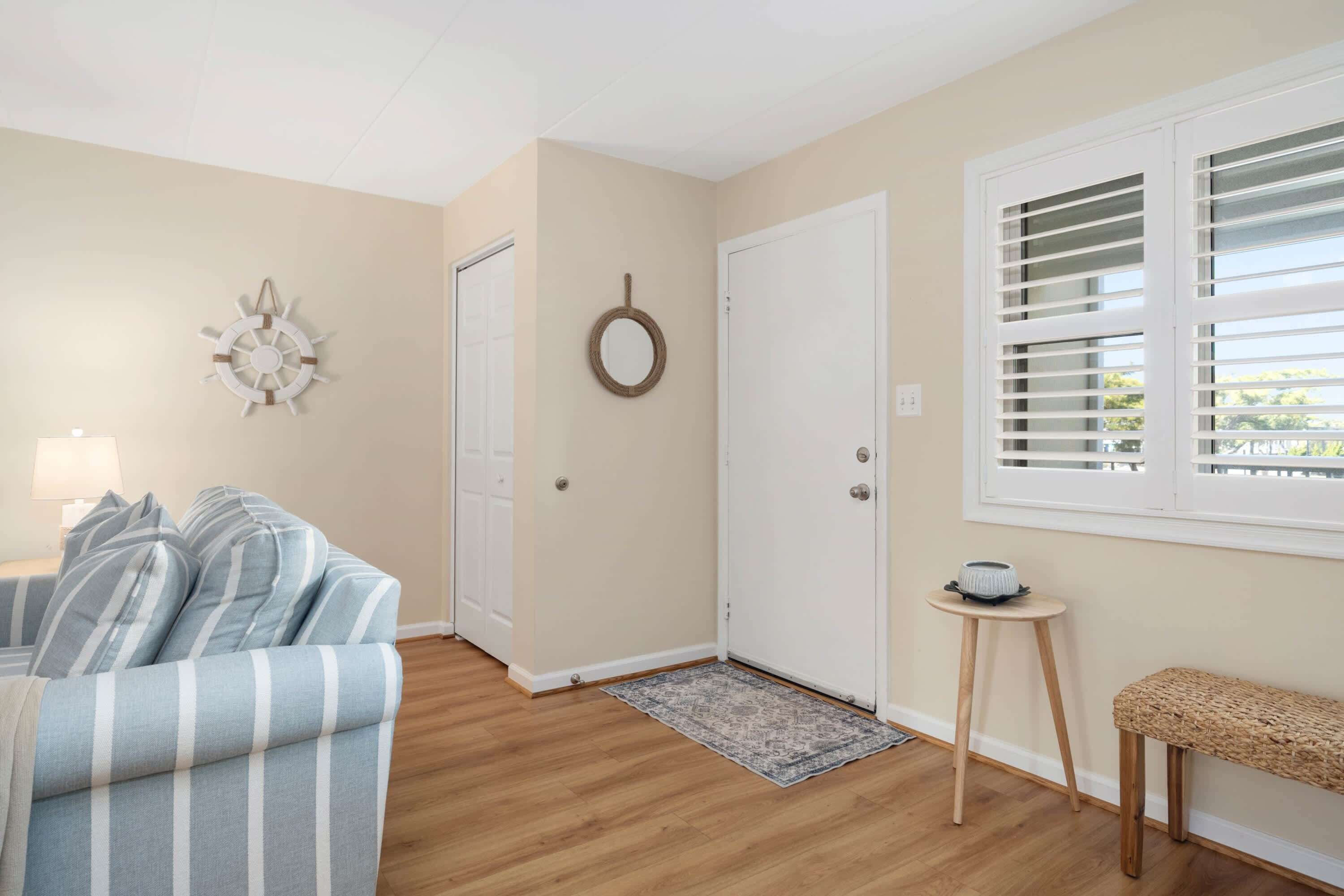 Una habitación con un sofá a rayas, una mesita redonda con un cuenco, un banco tejido, una puerta con un espejo y una ventana con persianas.