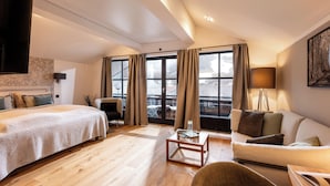 Een moderne hotelkamer met een groot bed, een bank, een bijzettafeltje en uitzicht op de stad door grote ramen.