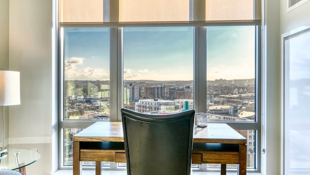 Un coin repas avec une grande fenêtre donnant sur la ville, une table, des chaises et une lampe.