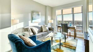 Un salon moderne avec un canapé, un fauteuil et une table basse en verre. Une grande fenêtre offre une vue sur la ville.