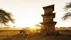 A rustic wooden building with multiple levels, a covered outdoor seating area, and a vehicle parked nearby. - Unique 2-story tower with 360° wildlife views and stargazing showers. (Naivasha)