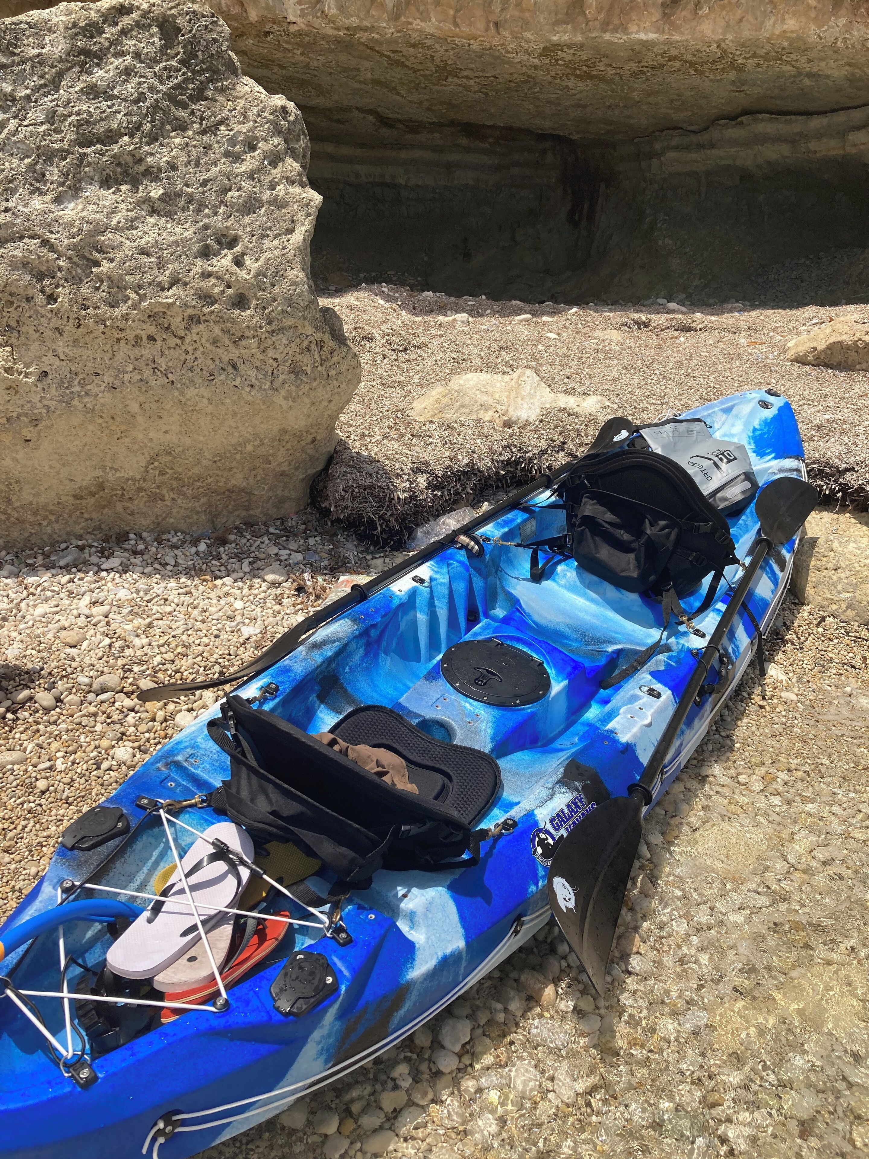 A blue kayak with a black bag and paddle, resting on a rocky surface near a cave entrance.