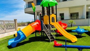 A playground with slides and climbing structures, surrounded by a well-maintained lawn and a building in the background. - Appartement à Louer Garden Paradise Bouznika (bouznika)