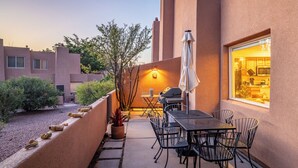 Une terrasse extérieure avec une table et des chaises, un barbecue et une vue sur la cuisine à travers une fenêtre.