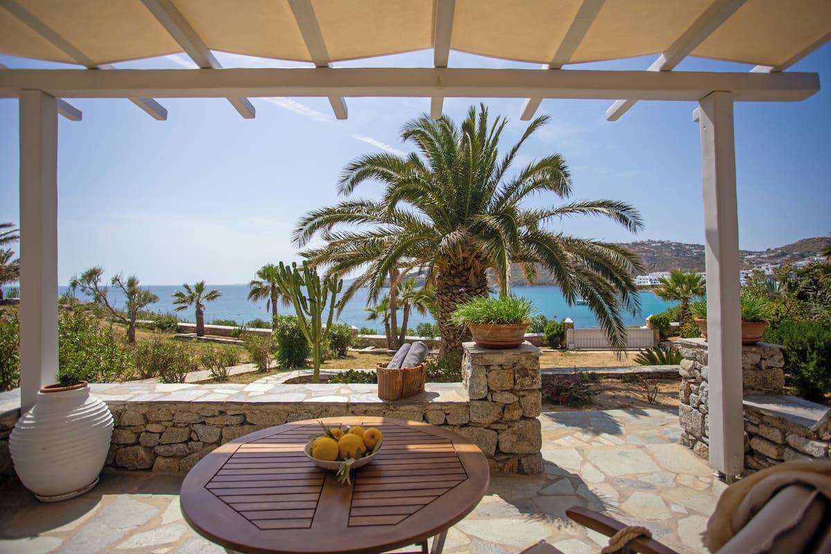 A covered outdoor seating area with a table and chairs, a bowl of lemons, and a view of the sea and palm trees.