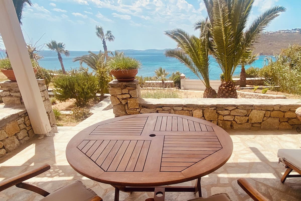 A covered outdoor seating area with a table and chairs, overlooking a scenic view of the sea and palm trees.