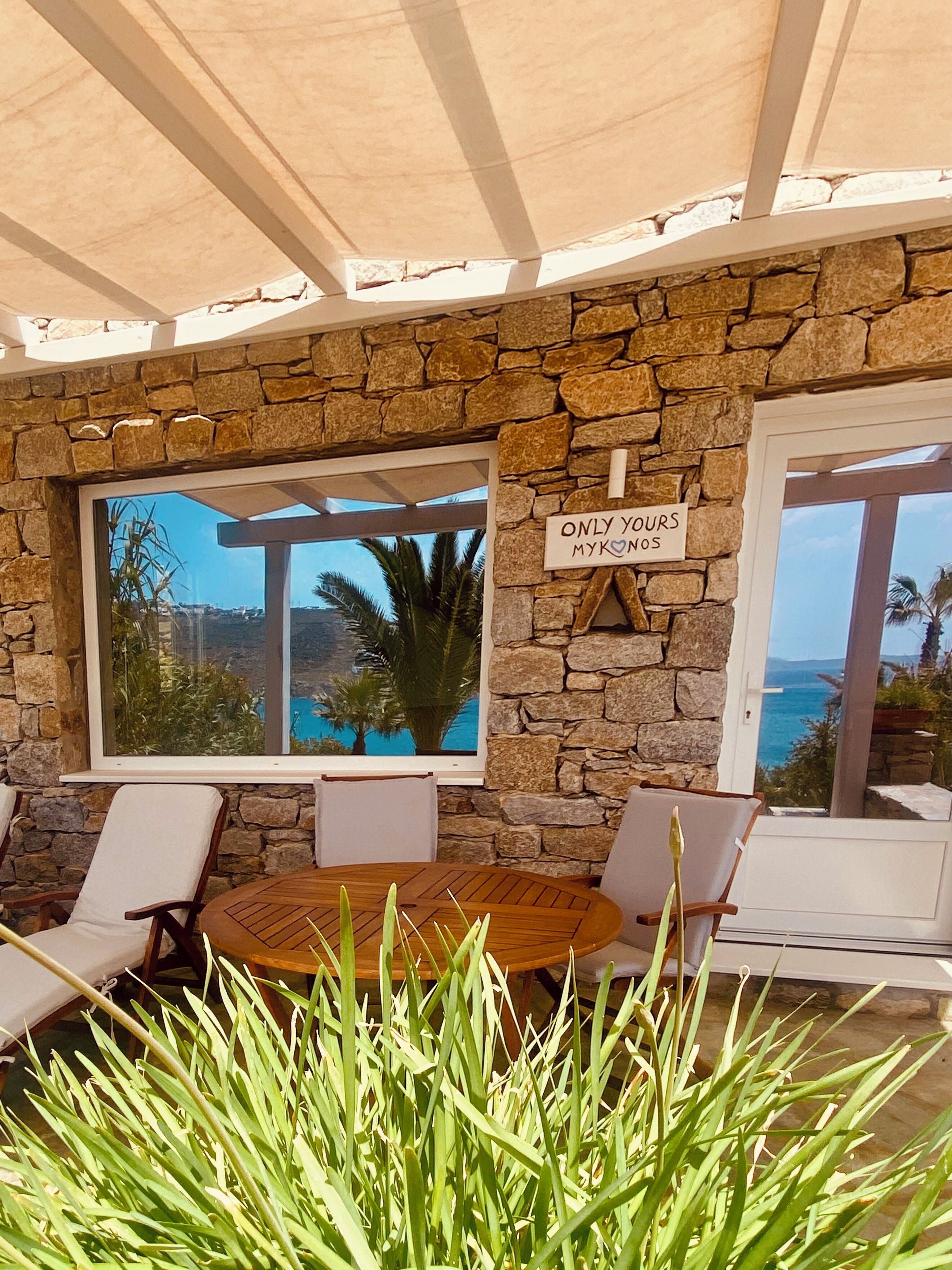 A stone wall with a sign reading "ONLY YOURS MYKONOS" and a view of the sea through large windows.
