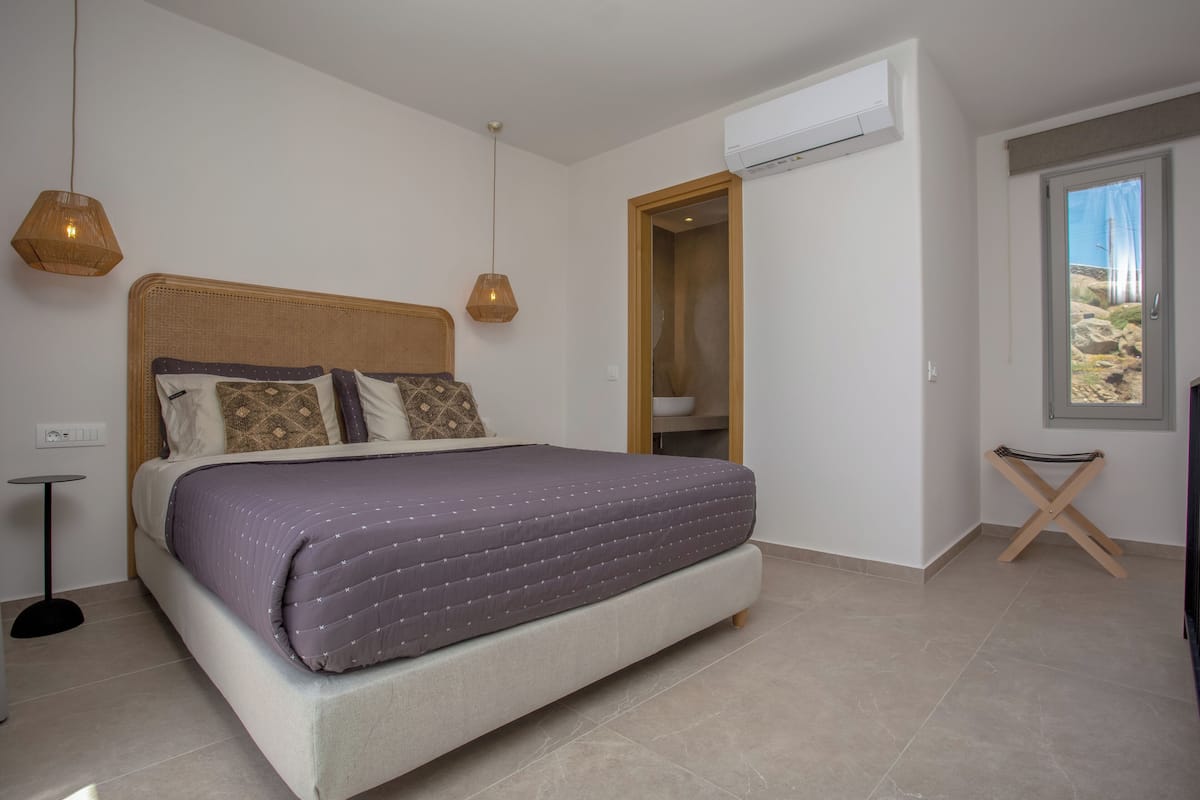 A bedroom with a bed, bedside lamps, an air conditioning unit, and a window with a view.