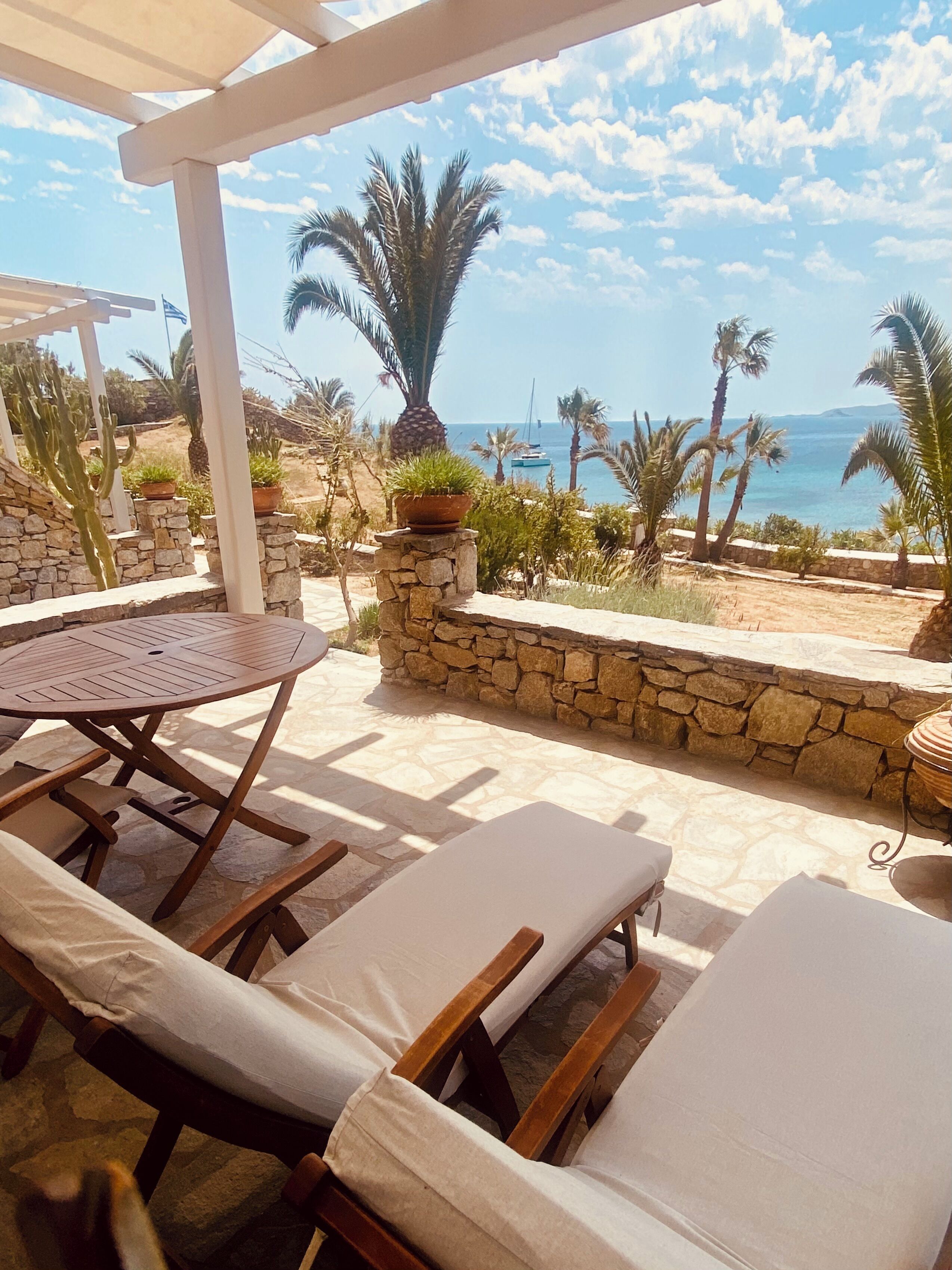 A patio with lounge chairs and a table, overlooking a beach with palm trees and boats.