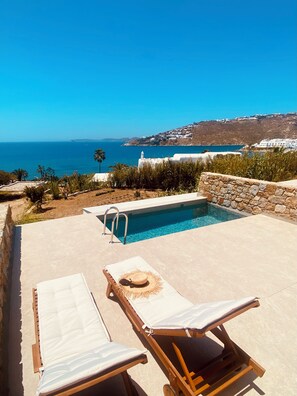 A poolside area with lounge chairs and a stone wall, overlooking a coastal landscape with a clear view of the sea and distant hills. - Only Yours Mykonos (Mykonos)