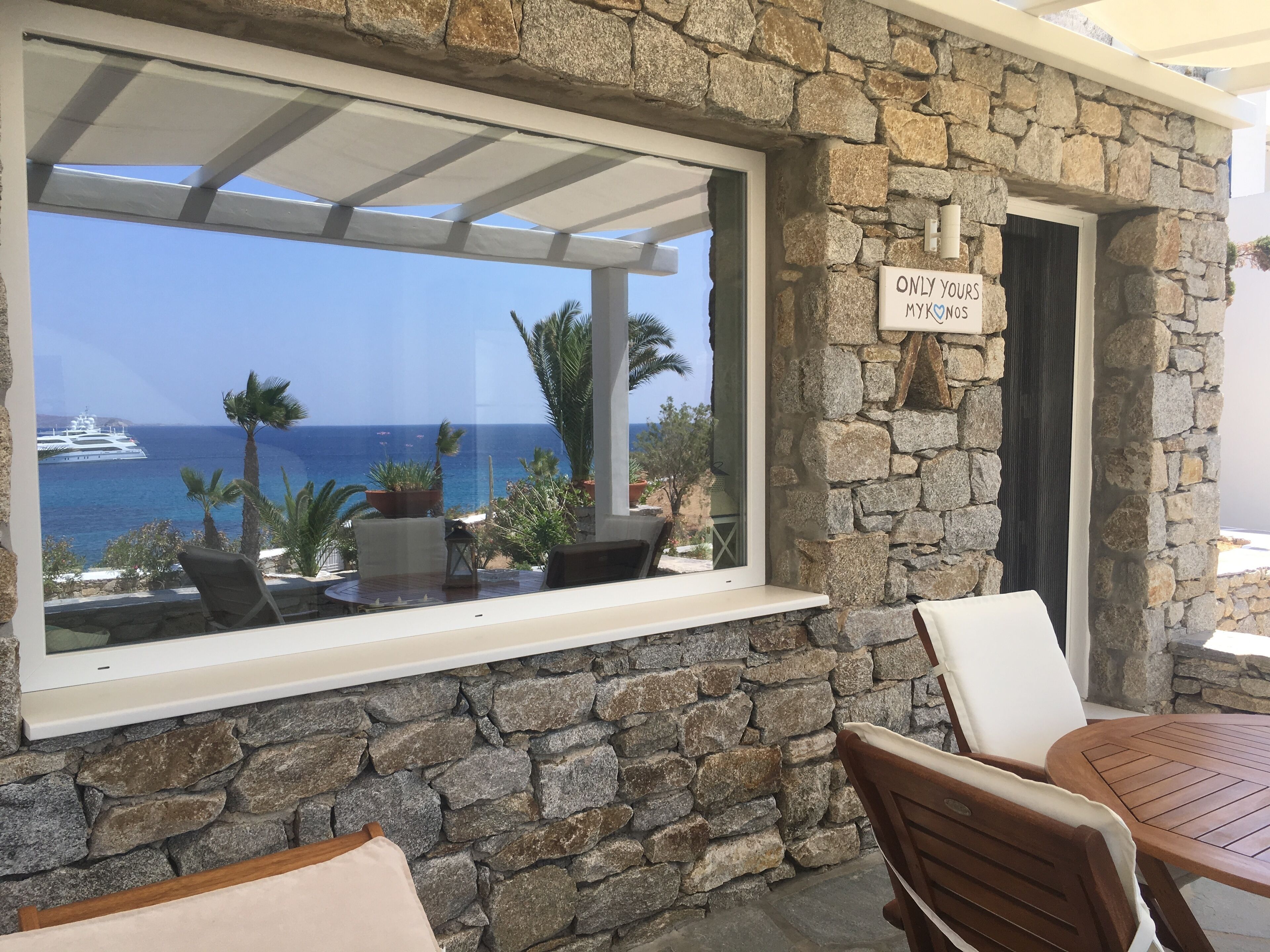 A stone wall with a window overlooking the sea, a sign reading "ONLY YOUR MYK NOS", and outdoor seating.