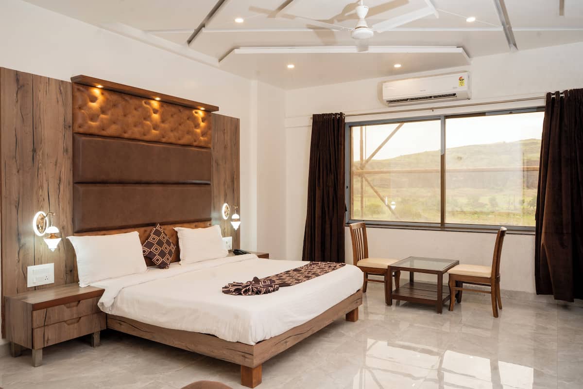 Quarto de hotel moderno com uma cama grande, uma mesinha e cadeiras, um ventilador de teto e uma janela ampla com cortinas.