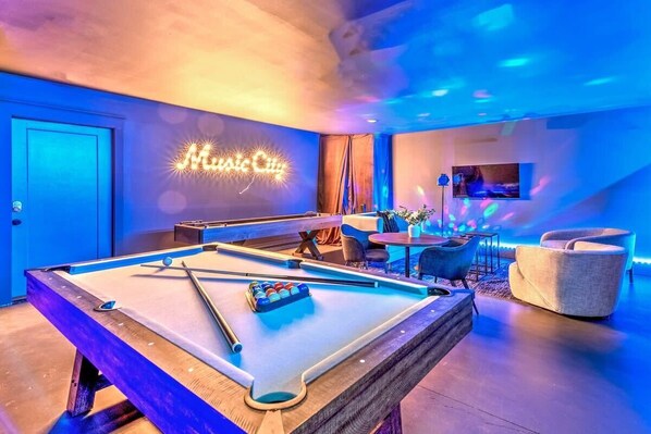 Une table de billard, surmontée d’un panneau néon indiquant « Music City », se trouve dans une pièce au design moderne, comprenant un bar et un espace détente avec des sièges.