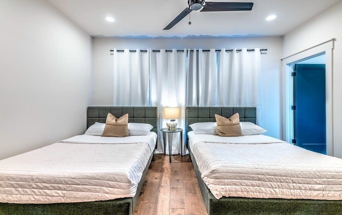 Habitación de hotel con dos camas, ventilador de techo, mesita de noche con lámpara y vista al exterior a través de una ventana con cortinas.
