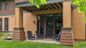 A covered patio area with two Adirondack chairs and a small table, adjacent to a building with large glass doors and windows.