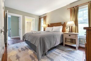Une chambre à coucher comprenant un lit, des lampes de chevet, une table de chevet, une fenêtre avec des rideaux et une porte donnant sur une autre pièce.