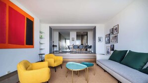 Een moderne woonkamer met een bank, twee gele stoelen, een ronde tafel en een eethoek op de achtergrond.