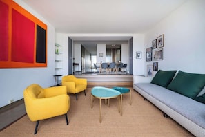 Ein modernes Wohnzimmer mit einer Couch, zwei gelben Sesseln, einem runden Tisch und einem Essbereich im Hintergrund.