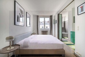 A hotel room with a bed, a bedside table, a lamp, a window with a view of buildings, and a door leading to a balcony. - Elegant Paris Apartment on Avenue Montaigne (Paris)