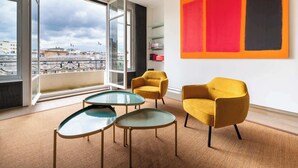 Een moderne kamer met een groot raam, twee gele fauteuils, een glazen salontafel en een abstract schilderij in rood en zwart.