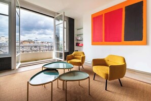 Ein modernes Zimmer mit einem großen Fenster, zwei gelben Sessel, einem Glastisch und einem abstraktem Gemälde in Rot und Schwarz.