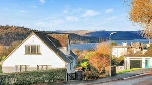 A white house with a gabled roof, a garage, and a view of a lake and hills in the background. - Beechwood Brook, 3 bed, 3 bath, parking, Bowness (Bowness-on-Windermere)