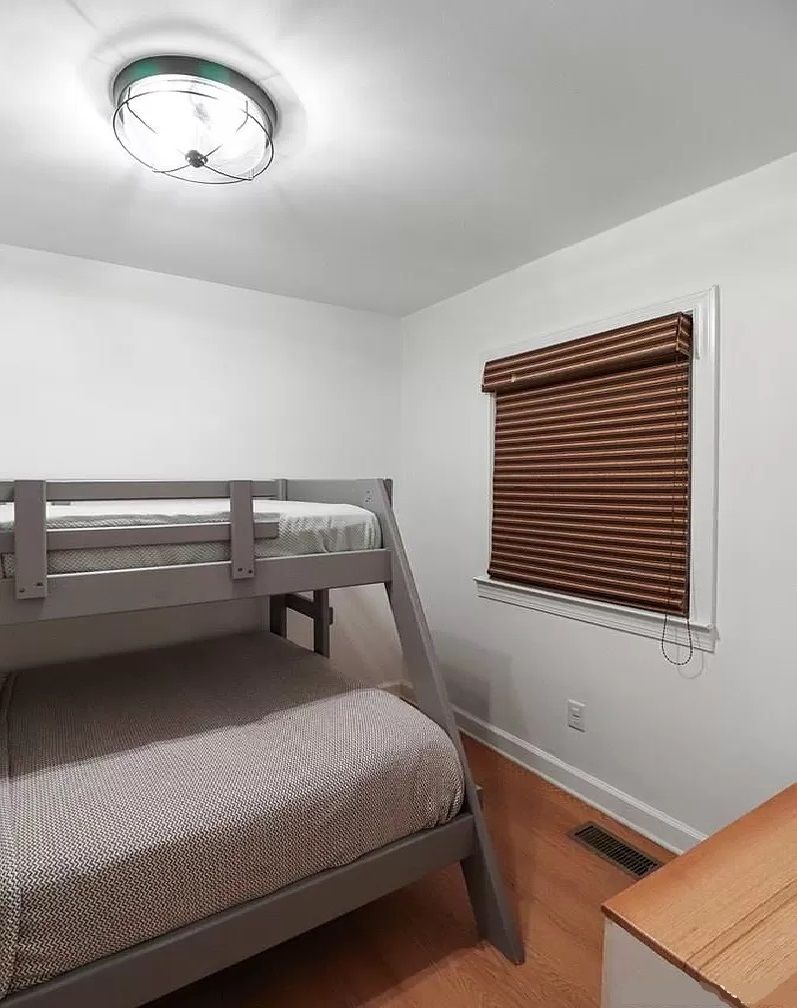 Een kleine kamer met een enkel bed en een stapelbed, een raam met zonwering en een plafondlamp.
