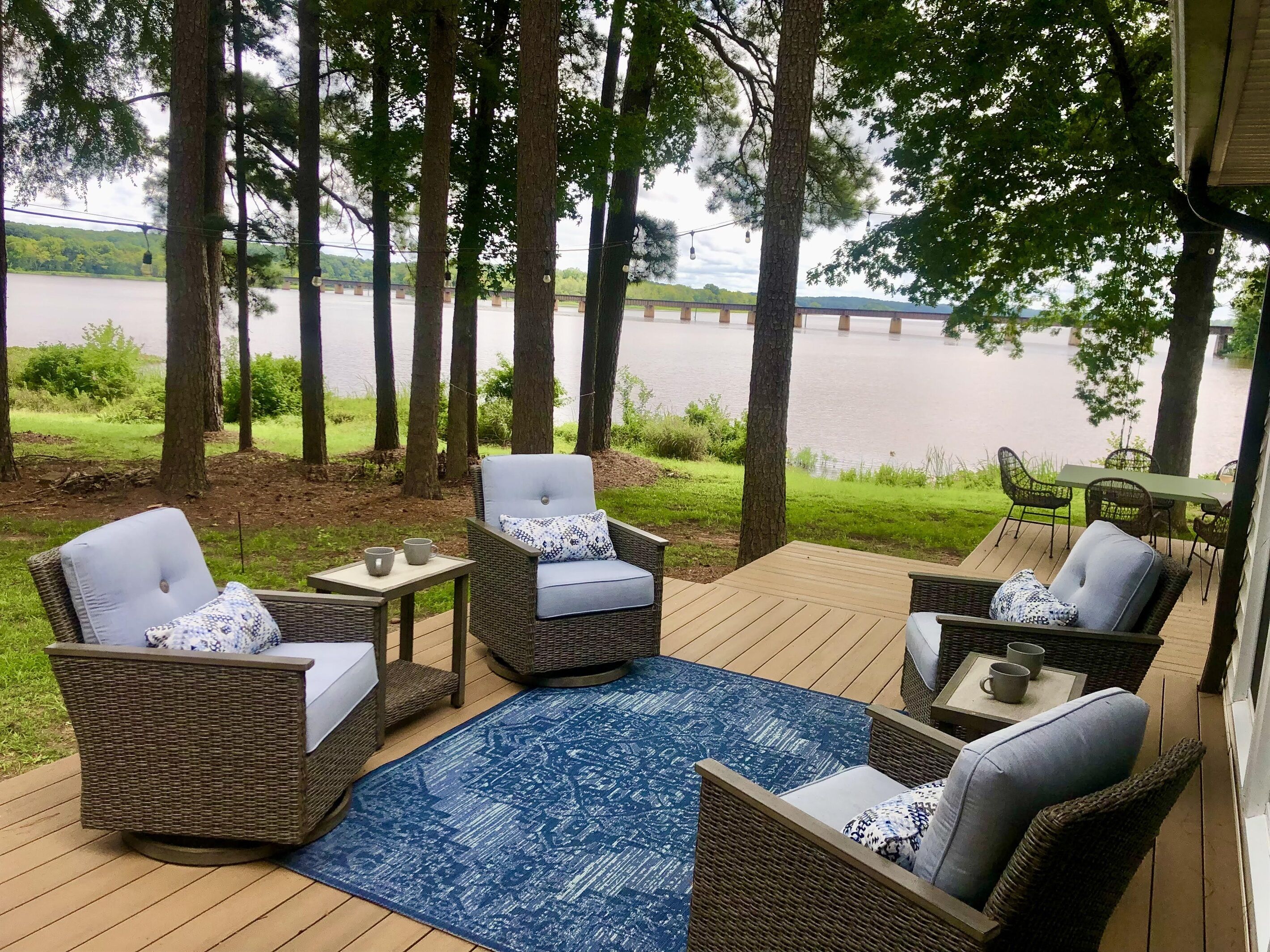 Una terraza con mobiliario de mimbre, una alfombra azul y vista a un lago y un puente.