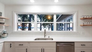 A kitchen with white cabinets, a stainless steel sink, and a dishwasher. There is a window above the sink with a view of trees and a building outside.