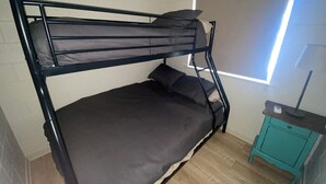 Una cama litera con colchón y almohadas, un pequeño mueble color turquesa y una ventana con persianas.