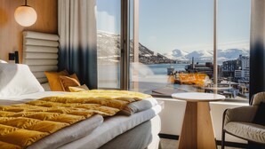 A modern hotel room with a large bed, a small round table, and a view of a harbor with boats and mountains. - The Dock 69°39 by Scandic (Tromsø)