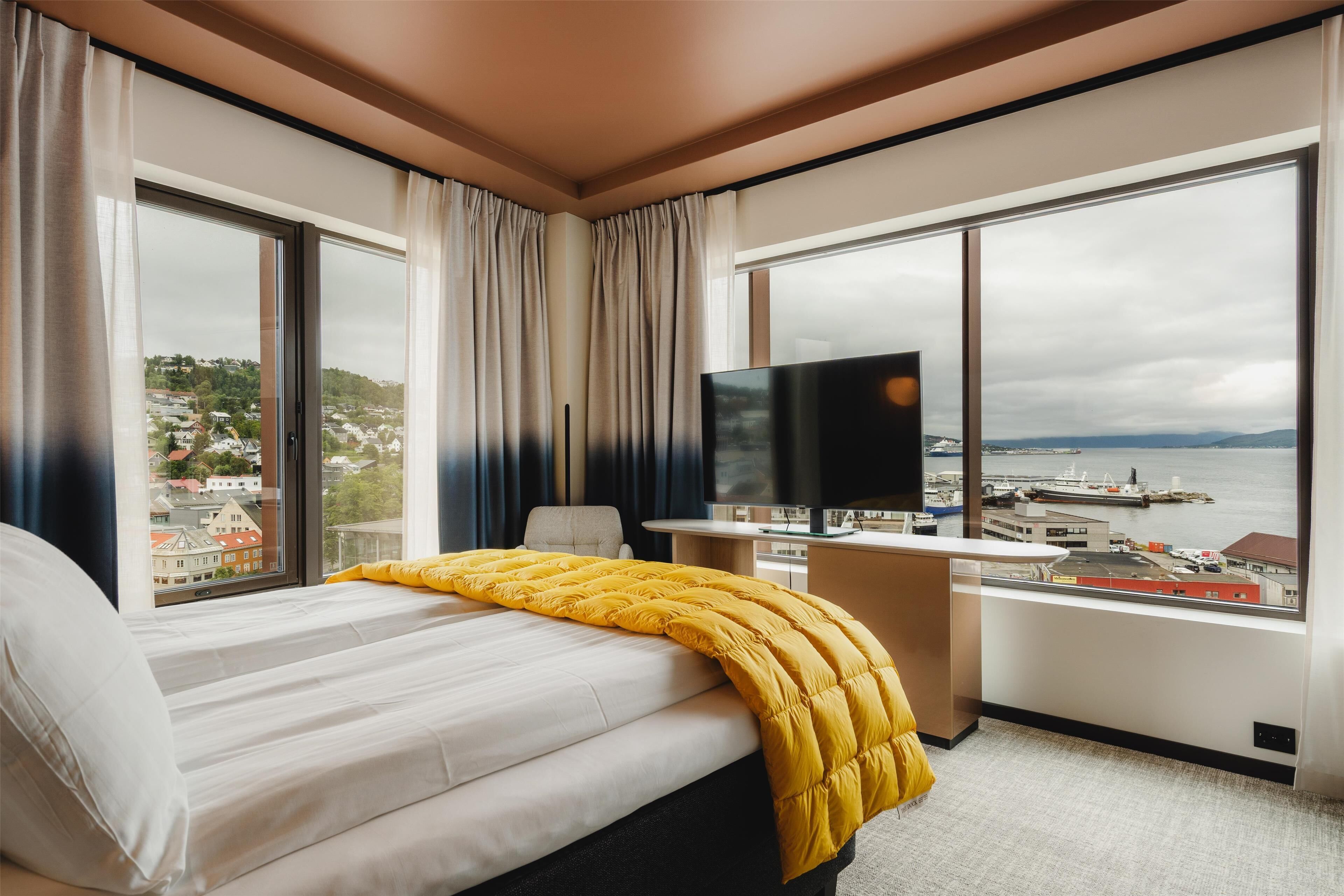 A hotel room with a bed, a desk, a TV, and large windows with a city and port view.