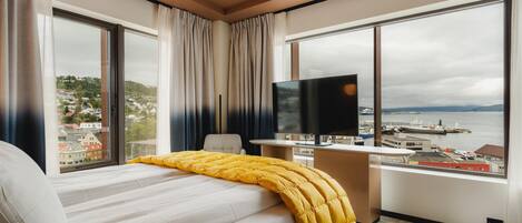 A hotel room with a bed, a desk, a TV, and large windows with a city and port view.