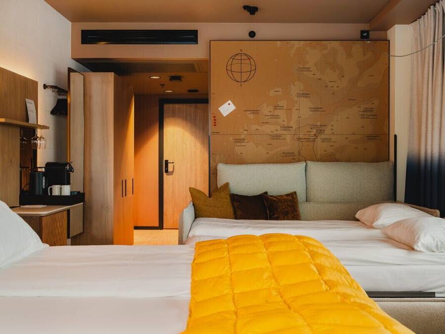 A hotel room with a bed, a desk with a coffee machine, a map on the wall, and a window with curtains.