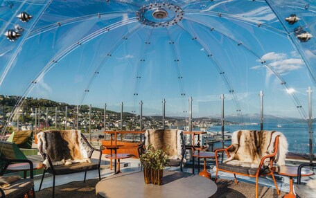 A transparent dome structure with seating and tables, overlooking a coastal cityscape.