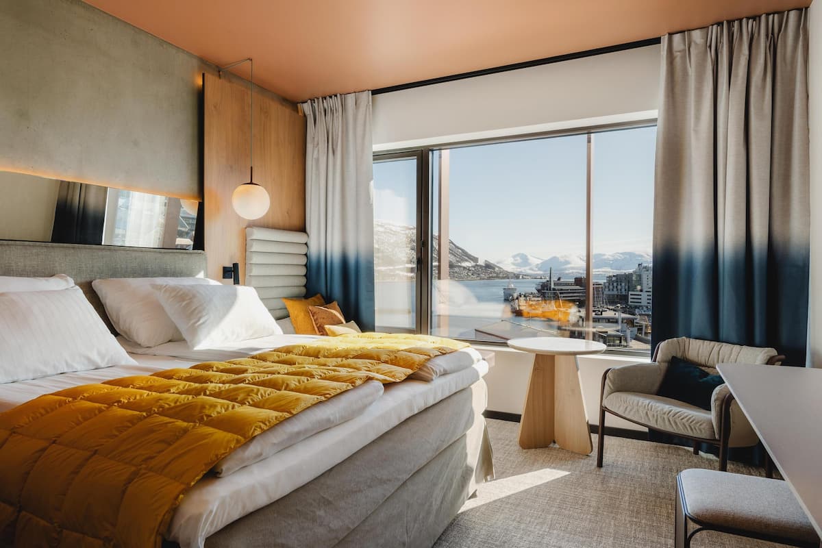 Une chambre d’hôtel moderne avec un grand lit, une petite table ronde, un canapé et une grande fenêtre donnant sur la ville.