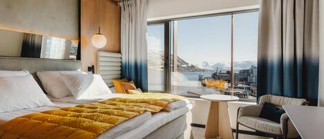 A modern hotel room with a large bed, a small round table, a sofa, and a large window offering a city view.