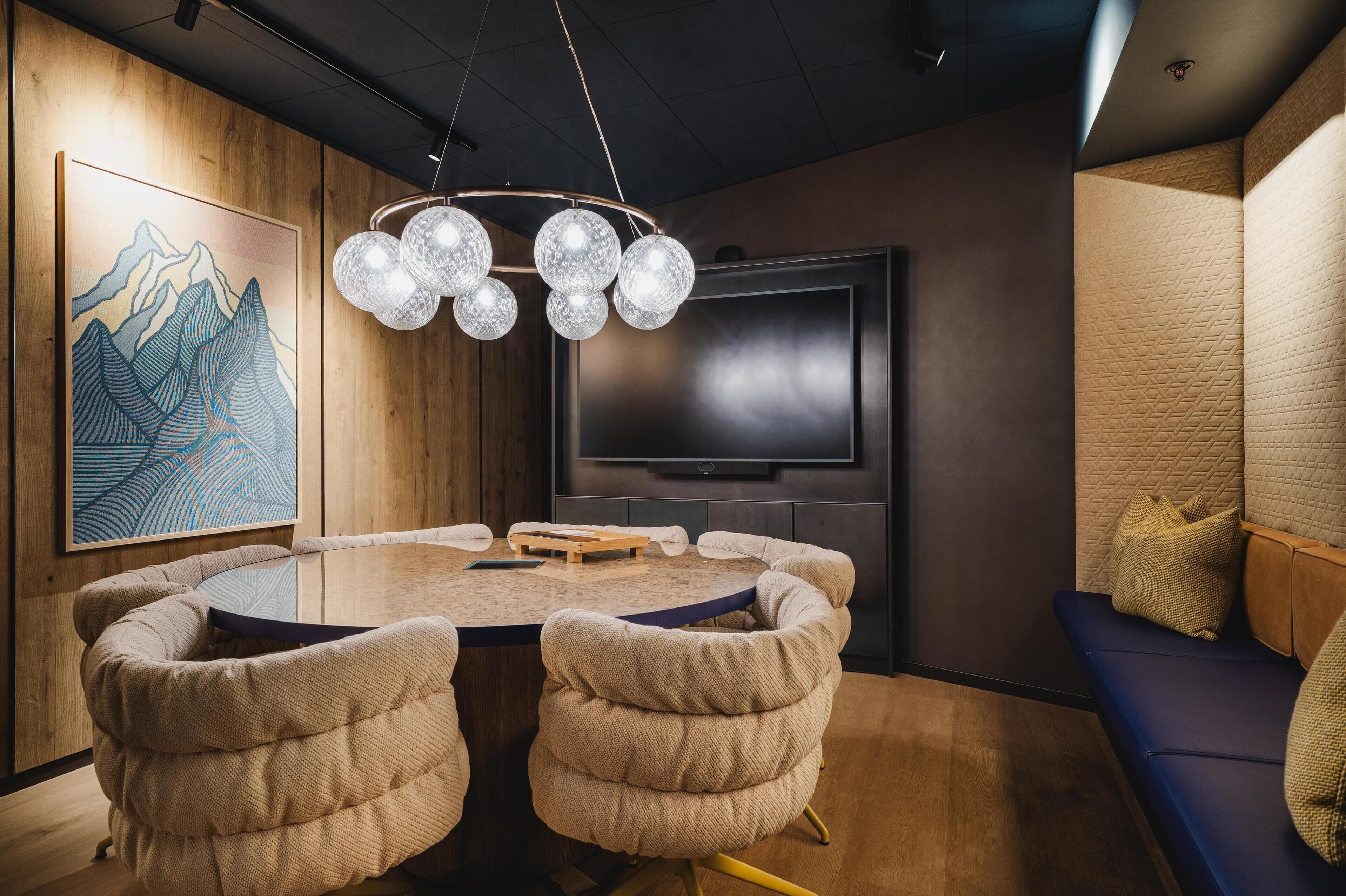 Une salle de conférence dotée d’une table ronde, de chaises beiges recouvertes de tissu, d’un téléviseur fixé au mur et d’un mur en bois orné d’un tableau représentant une montagne.
