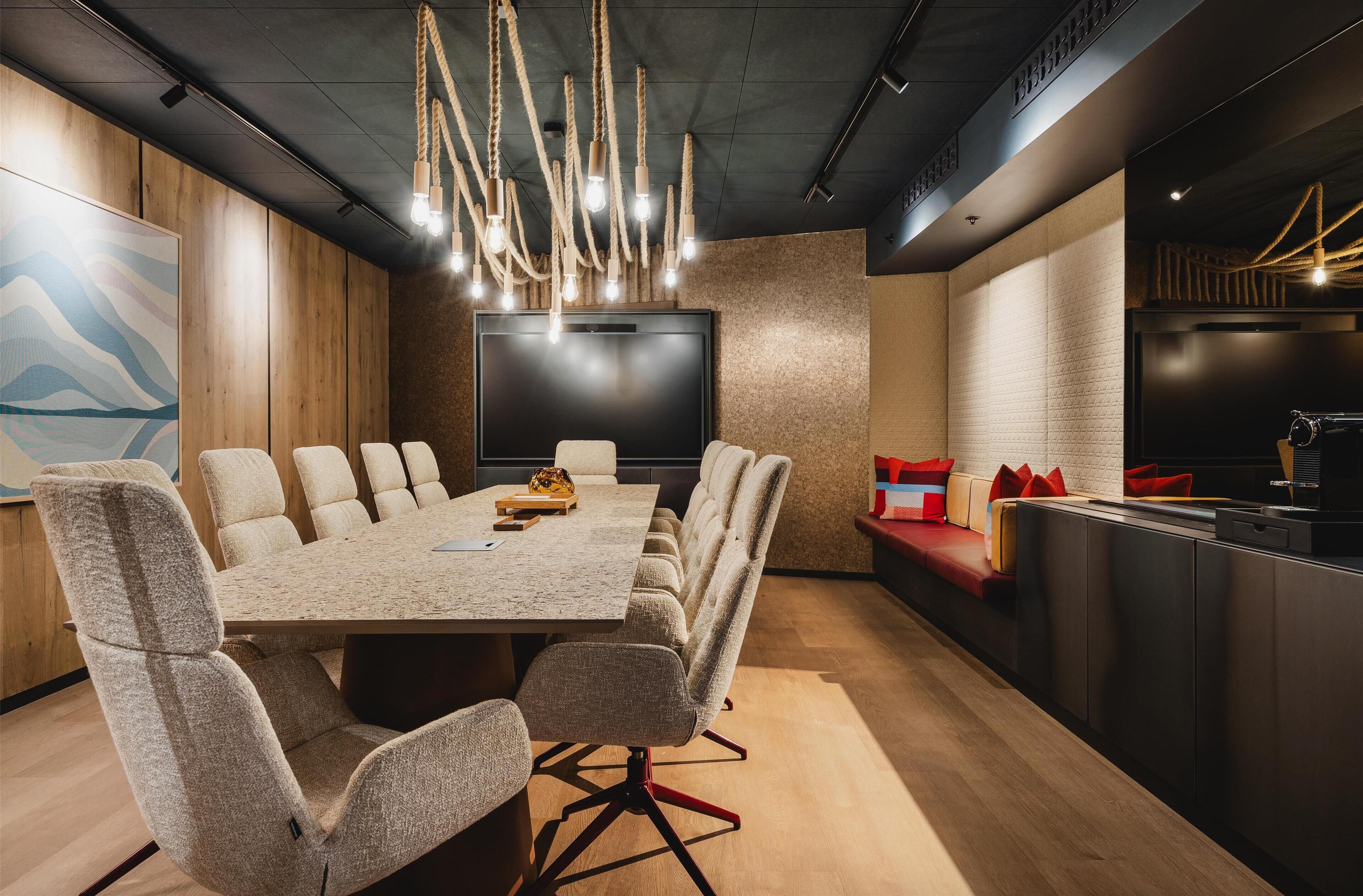 A modern conference room with a long table, comfortable chairs, a large screen, and a kitchen area with seating.