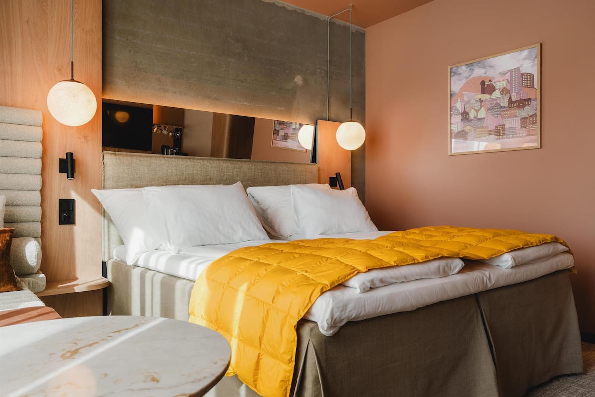 Un lit bien fait, avec une couverture jaune, une tête de lit en bois et une œuvre d’art encadrée représentant un paysage urbain accrochée au mur.
