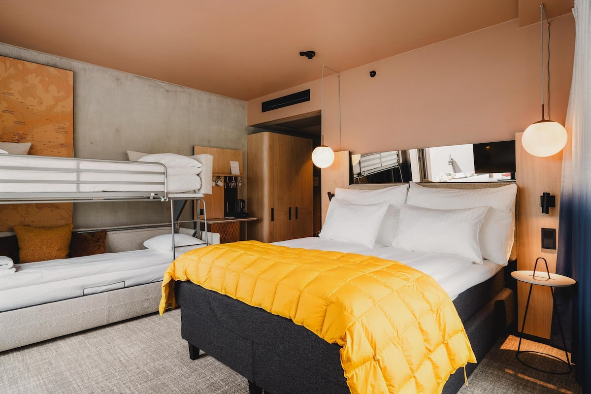 Une chambre d’hôtel avec un lit superposé, un lit simple recouvert d’une couverture jaune, une petite table et un luminaire suspendu.