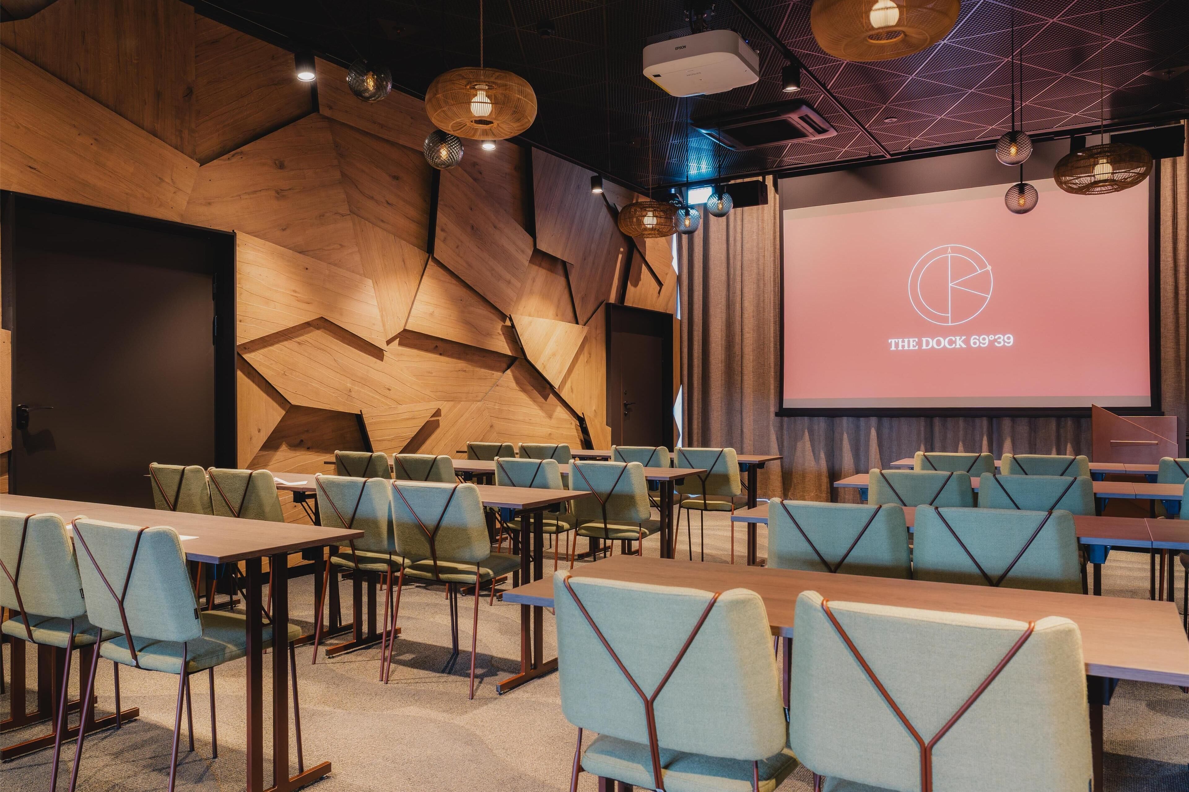 Una sala de conferencias con una pantalla grande mostrando 'THE DOCK 69P39', varias mesas y sillas dispuestas para una reunión.