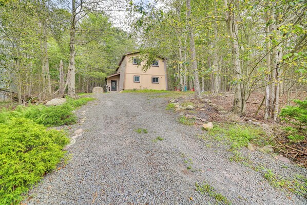 Une maison à deux étages, avec une allée en gravier menant à une porte en bois, entourée d’arbres denses et d’une végétation luxuriante.